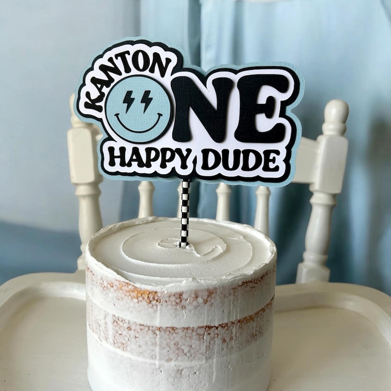 May include: A blue and white cake topper with the text "Kanton One Happy Dude" and a lightning bolt smiley face. The topper is on a white cake.