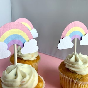 May include: Three cupcakes topped with white frosting and rainbow-coloured sprinkles. Each cupcake has a paper rainbow and cloud topper.