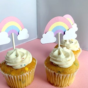May include: Four cupcakes with white frosting and colourful sprinkles topped with rainbow cupcake toppers. The toppers are made of paper and feature a pink, yellow, and purple rainbow with white clouds.