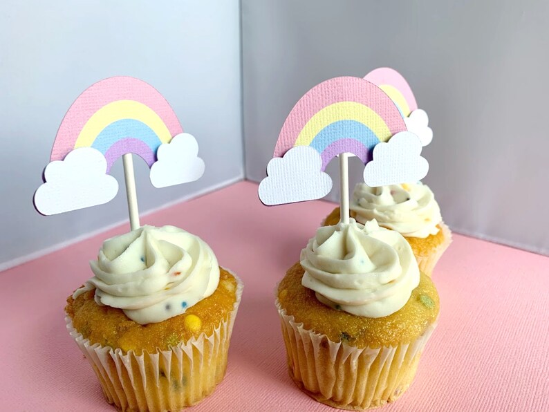 May include: Four cupcakes with white frosting and colourful sprinkles topped with rainbow cupcake toppers. The toppers are made of paper and feature a pink, yellow, and purple rainbow with white clouds.