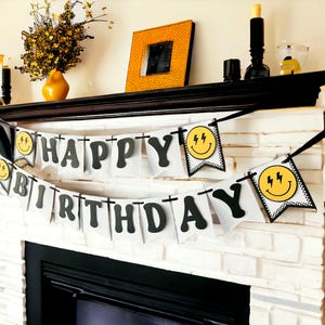 May include: A white banner with black letters spelling "HAPPY BIRTHDAY" hangs in front of a white brick fireplace. The banner includes smiley face flags with lightning bolt accents. A yellow vase and picture frame are on the mantel.