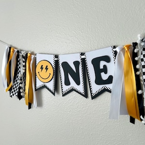 May include: A festive banner with the word "ONE" in black letters on white pennants. The banner features a smiley face with lightning bolts and is decorated with yellow, white, and black ribbons and checkered flags. Perfect for a first birthday celebration.