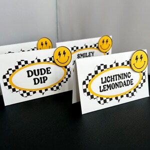 May include: White cardstock place cards with black and yellow checkered borders and text. Each card has a yellow smiley face with lightning bolts. The text on the cards reads "DUDE DIP", "SMILEY", and "LIGHTNING LEMONADE".
