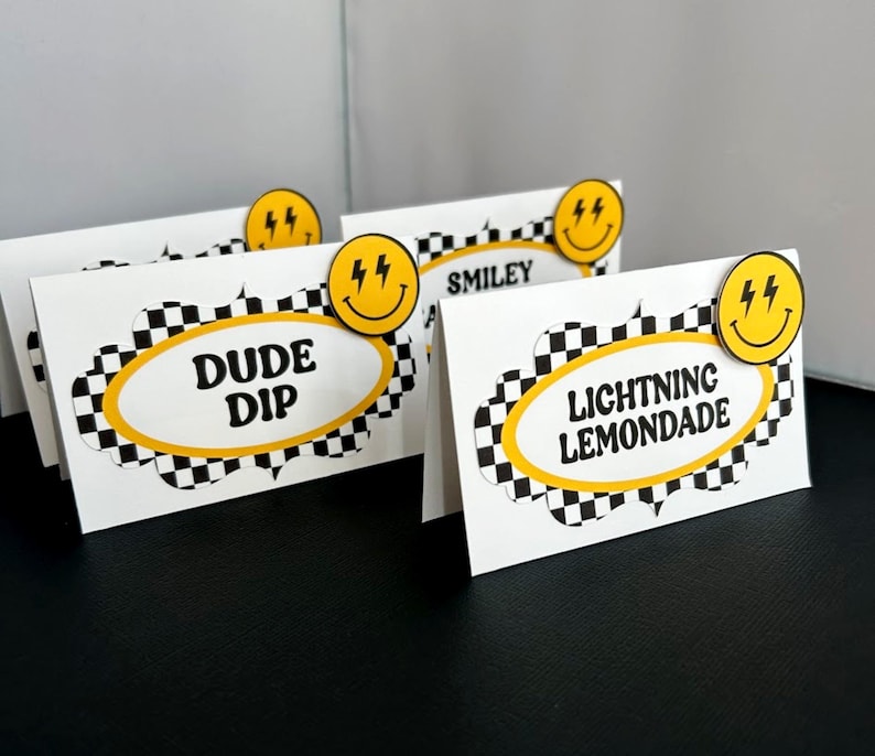 May include: White cardstock place cards with black and yellow checkered borders and text. Each card has a yellow smiley face with lightning bolts. The text on the cards reads "DUDE DIP", "SMILEY", and "LIGHTNING LEMONADE".