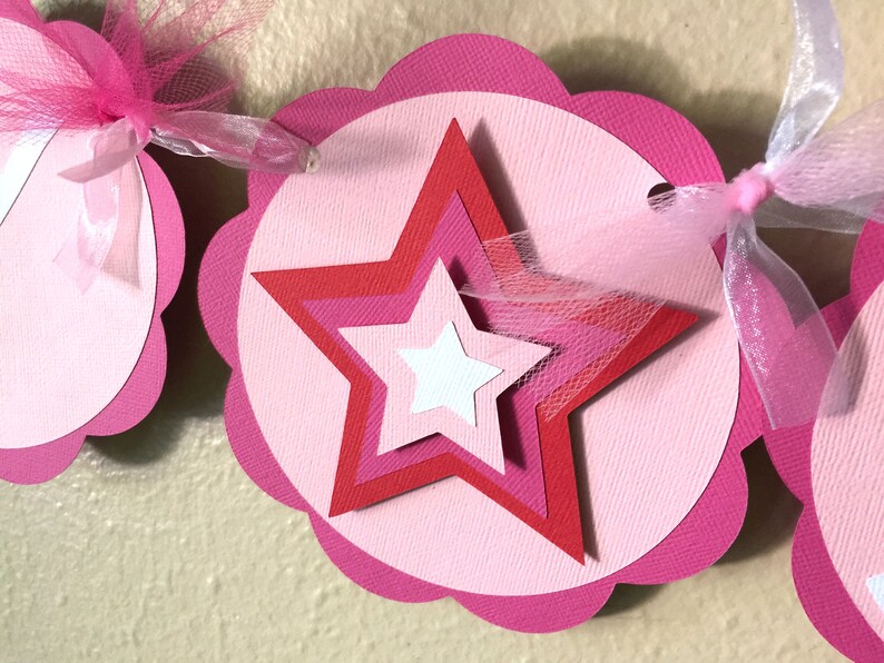 May include: Pink and red paper star banner with pink tulle ribbon. The banner is made of scalloped circles with a star in the centre.