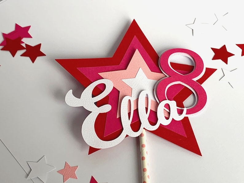 May include: A pink and red layered star cake topper with the name "Ella" and the number "8" in white. The topper is on a white background with small pink and white stars.