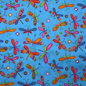May include: A blue fabric with a repeating pattern of colorful dragonflies. The dragonflies have striped wings in shades of red, orange, pink, and green, with floral and patterned details. Small floral and star accents are scattered throughout.