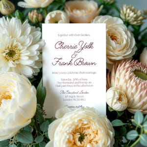 May include: A wedding invitation featuring the names "Cherrie Yolk & Frank Brown" in a stylish font. The invitation is set against a backdrop of cream and white flowers. The text includes the date, time, and venue in London.
