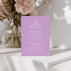 May include: A lilac wedding invitation card with the text "The Wedding Weekend." Details include a welcome party and farewell brunch with dates, times, and locations. A vase of flowers is in the background.