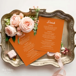 May include: Two orange wedding details cards with white script on a silver tray, decorated with peach roses and greenery. A small pink box and a few yellow gems are also present. The cards read "Details".
