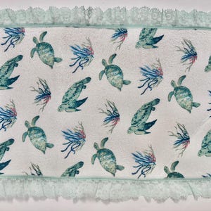 May include: White rectangular towel with a repeating pattern of teal sea turtles and blue and red sea plants. The towel has a light green lace trim around the edges.