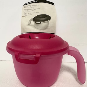May include: A pink Tupperware Mini Rice Maker with a handle and a lid. The product has a locking mechanism and a label with the Tupperware logo and product information.
