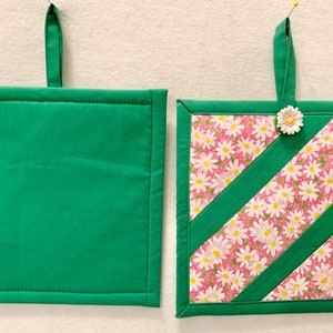 Puede incluir: Dos agarraderas cuadradas con lazos de tela verde. Una es de color verde s&oacute;lido, mientras que la otra presenta un fondo rosa con un patr&oacute;n floral de margaritas blancas, acentuado por dos tiras diagonales de tela verde y una peque&ntilde;a flor.