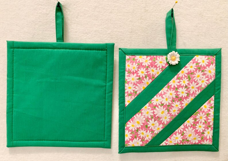 Puede incluir: Dos agarraderas cuadradas con lazos de tela verde. Una es de color verde s&oacute;lido, mientras que la otra presenta un fondo rosa con un patr&oacute;n floral de margaritas blancas, acentuado por dos tiras diagonales de tela verde y una peque&ntilde;a flor.