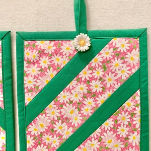 Puede incluir: Un agarrador de cocina cuadrado acolchado con tela rosa y estampado de margaritas blancas, acentuado por dos tiras diagonales de tela verde s&oacute;lida. El borde es verde y un peque&ntilde;o adorno de margarita blanca est&aacute; unido al lazo superior.