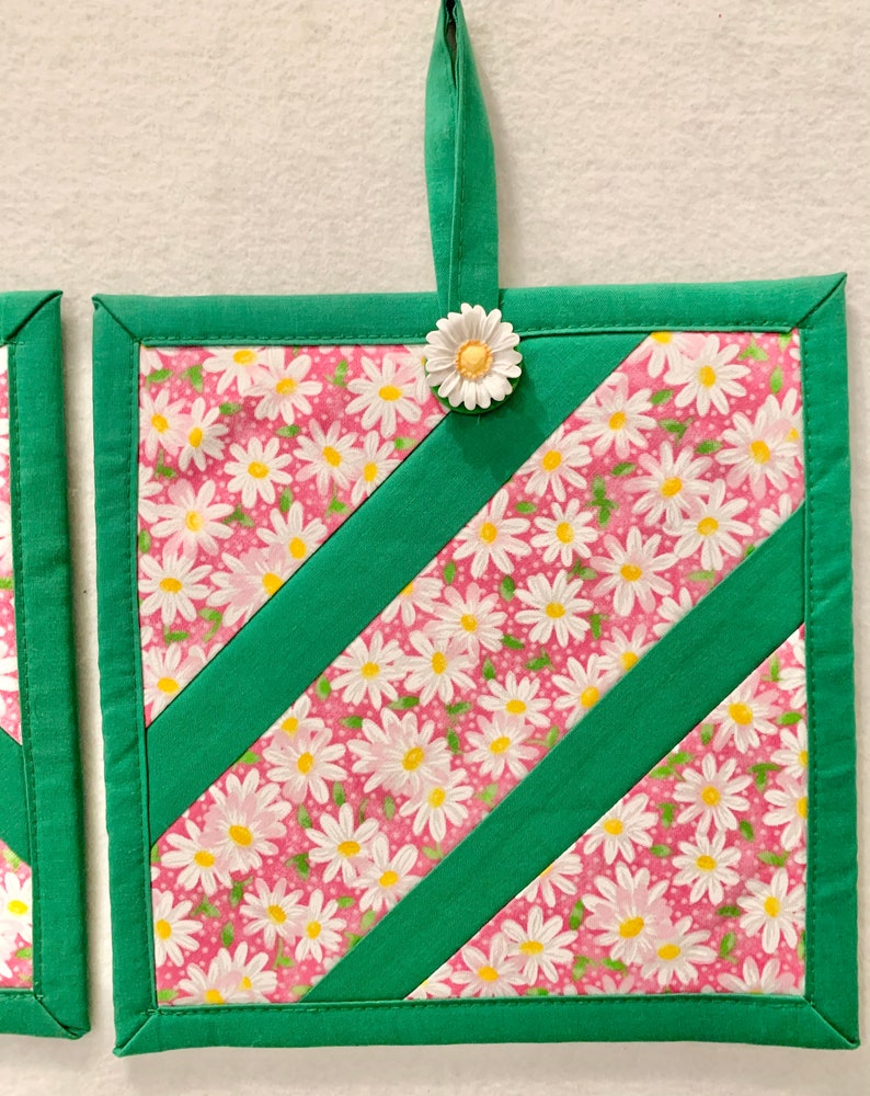 Puede incluir: Un agarrador de cocina cuadrado acolchado con tela rosa y estampado de margaritas blancas, acentuado por dos tiras diagonales de tela verde s&oacute;lida. El borde es verde y un peque&ntilde;o adorno de margarita blanca est&aacute; unido al lazo superior.