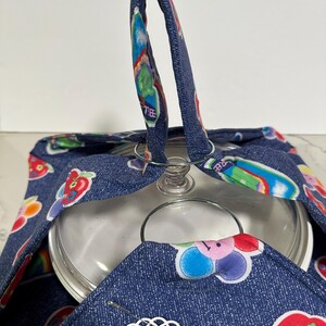 May include: A denim casserole carrier with a colorful fruit and flower pattern. The carrier has a handle and a decorative tag that reads "Hot or Cold Casserole Carrier". A glass lid is visible inside the carrier.