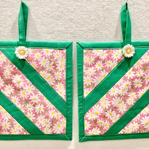 Puede incluir: Dos agarraderas de tela cuadradas con fondo rosa y estampado floral de margaritas blancas, acentuadas por tiras de tela diagonales y bordes en verde s&oacute;lido. Cada agarradera incluye una presilla de tela verde y un adorno de margarita blanca.