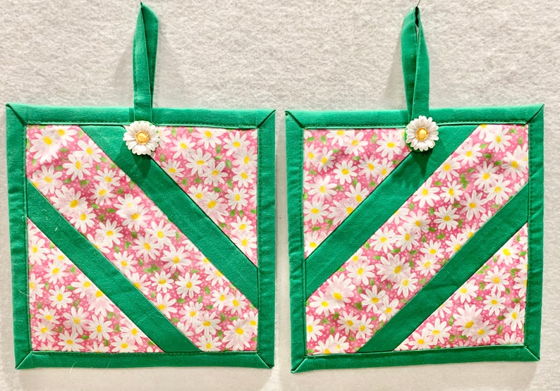 Puede incluir: Dos agarraderas de tela cuadradas con fondo rosa y estampado floral de margaritas blancas, acentuadas por tiras de tela diagonales y bordes en verde s&oacute;lido. Cada agarradera incluye una presilla de tela verde y un adorno de margarita blanca.