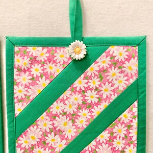 Puede incluir: Un agarrador de tela cuadrado con un fondo rosa y un patr&oacute;n floral de margaritas blancas, acentuado por dos tiras diagonales de tela verde s&oacute;lida. Los bordes est&aacute;n ribeteados en tela verde y una peque&ntilde;a margarita blanca adorna la parte superior.