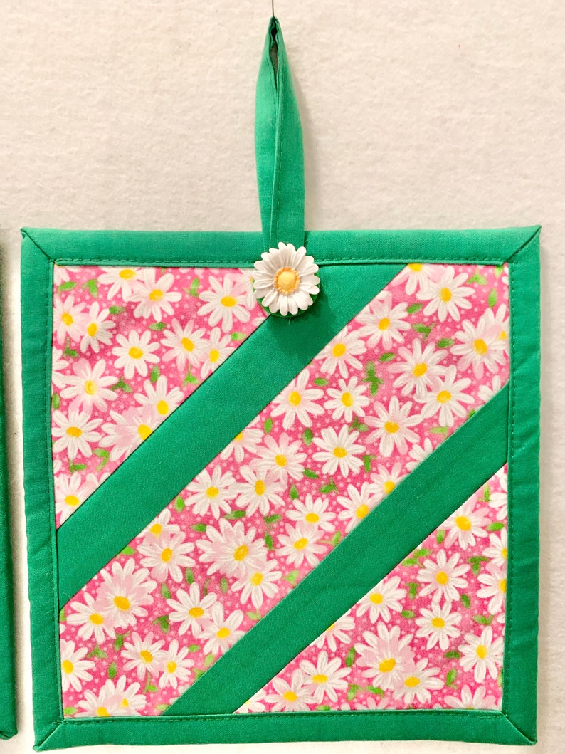 Puede incluir: Un agarrador de tela cuadrado con un fondo rosa y un patr&oacute;n floral de margaritas blancas, acentuado por dos tiras diagonales de tela verde s&oacute;lida. Los bordes est&aacute;n ribeteados en tela verde y una peque&ntilde;a margarita blanca adorna la parte superior.
