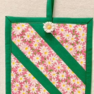 Puede incluir: Un agarrador de tela cuadrado con fondo rosa y estampado de margaritas blancas, acentuado por bordes y tiras diagonales de tela verde s&oacute;lida. Un bot&oacute;n en forma de margarita blanca est&aacute; sujeto en la parte superior central, con una presilla.