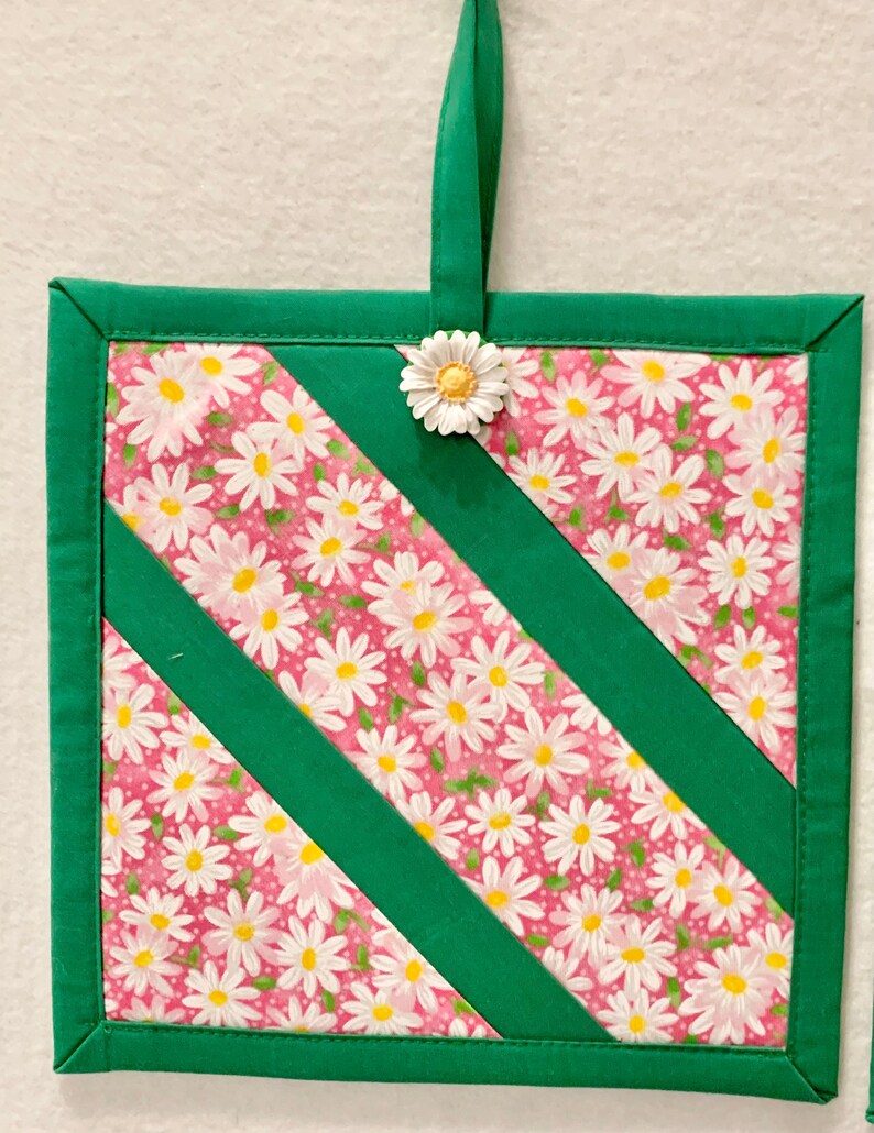 Puede incluir: Un agarrador de tela cuadrado con fondo rosa y estampado de margaritas blancas, acentuado por bordes y tiras diagonales de tela verde s&oacute;lida. Un bot&oacute;n en forma de margarita blanca est&aacute; sujeto en la parte superior central, con una presilla.