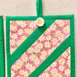 Puede incluir: Un agarrador de tela cuadrado con fondo rosa y estampado floral de margaritas blancas, acentuado por dos tiras de tela verde en diagonal. El borde es verde y un peque&ntilde;o adorno de margarita blanca est&aacute; sujeto al lazo para colgar.