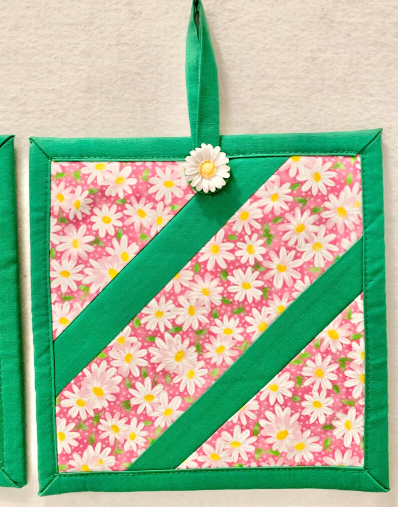 Puede incluir: Un agarrador de tela cuadrado con fondo rosa y estampado floral de margaritas blancas, acentuado por dos tiras de tela verde en diagonal. El borde es verde y un peque&ntilde;o adorno de margarita blanca est&aacute; sujeto al lazo para colgar.