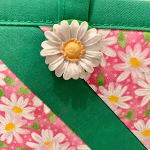 Puede incluir: Un art&iacute;culo de tela con fondo rosa y estampado floral de margaritas blancas, acentuado por bordes de tela verde s&oacute;lido y un lazo de tela. Una margarita blanca tridimensional con centro amarillo est&aacute; unida al borde verde.