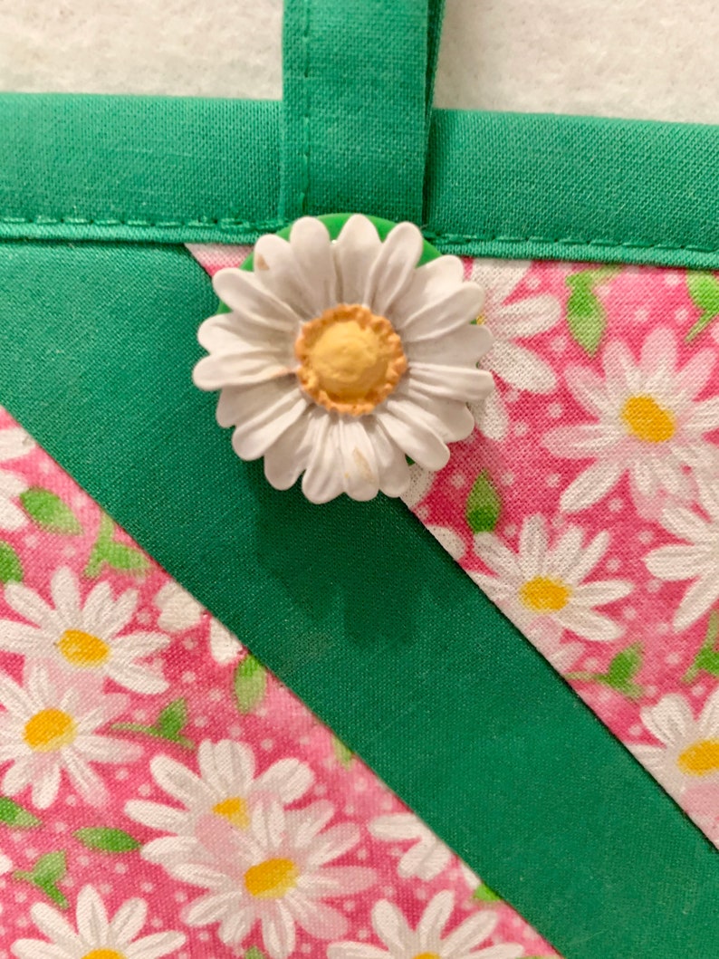 Puede incluir: Un art&iacute;culo de tela con fondo rosa y estampado floral de margaritas blancas, acentuado por bordes de tela verde s&oacute;lido y un lazo de tela. Una margarita blanca tridimensional con centro amarillo est&aacute; unida al borde verde.
