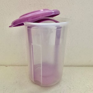 May include: A clear plastic container with a purple lid. The lid is partially open, revealing the interior of the container. The container is cylindrical with a slightly flared top. The overall design is simple and functional.