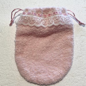 May include: A soft, pink drawstring bag with a textured surface. The top edge is adorned with white lace and pink ribbon accents. The bag is designed with a rounded bottom and a drawstring closure.