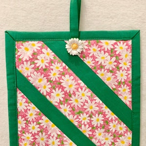 Puede incluir: Un agarrador de tela cuadrado con un patr&oacute;n floral de margaritas blancas sobre fondo rosa, acentuado por franjas diagonales de tela verde s&oacute;lida y un borde verde. Un adorno de margarita blanca est&aacute; sujeto al lazo central superior.