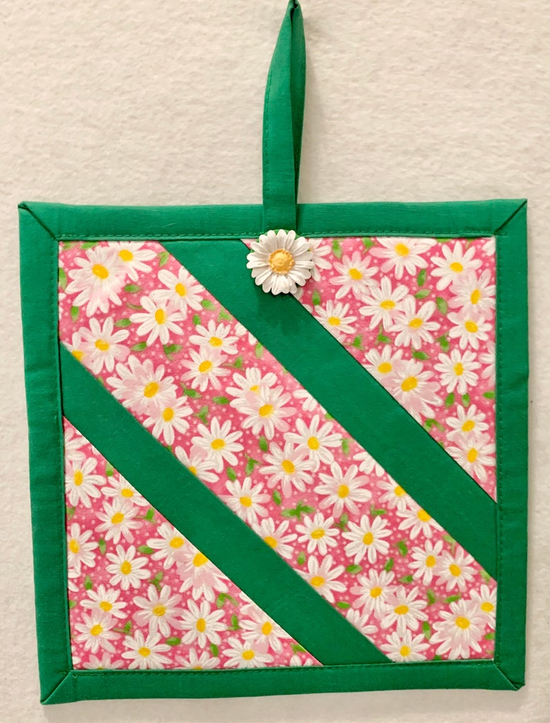 Puede incluir: Un agarrador de tela cuadrado con un patr&oacute;n floral de margaritas blancas sobre fondo rosa, acentuado por franjas diagonales de tela verde s&oacute;lida y un borde verde. Un adorno de margarita blanca est&aacute; sujeto al lazo central superior.