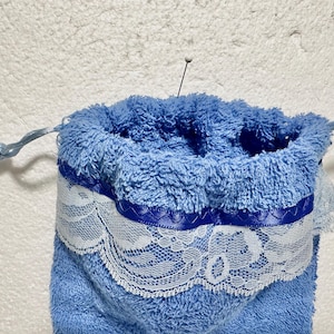 May include: A blue drawstring bag made of soft, fluffy material. The bag is trimmed with a white lace band and a blue ribbon. The drawstring is a light blue cord. A pin is inserted into the top of the bag.