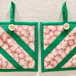 Puede incluir: Dos agarraderas cuadradas acolchadas de tela rosa con estampado de margaritas blancas, acentuadas con tiras de tela verde s&oacute;lida en diagonal y bordes verdes. Cada agarradera incluye un lazo de tela verde para colgar con un bot&oacute;n de margarita.
