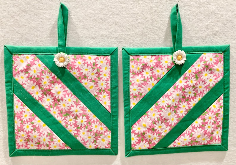Puede incluir: Dos agarraderas cuadradas acolchadas de tela rosa con estampado de margaritas blancas, acentuadas con tiras de tela verde s&oacute;lida en diagonal y bordes verdes. Cada agarradera incluye un lazo de tela verde para colgar con un bot&oacute;n de margarita.