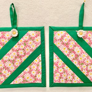 Puede incluir: Dos agarradores de tela cuadrados con fondo rosa y estampado floral de margaritas blancas, acentuados por tiras de tela diagonales en verde s&oacute;lido. Cada agarrador incluye una cinta de tela verde para colgar y un adorno de margarita.