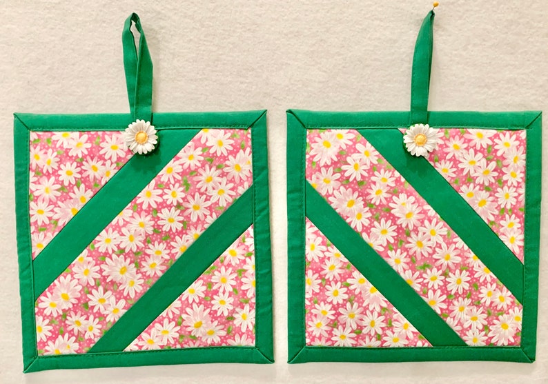 Puede incluir: Dos agarradores de tela cuadrados con fondo rosa y estampado floral de margaritas blancas, acentuados por tiras de tela diagonales en verde s&oacute;lido. Cada agarrador incluye una cinta de tela verde para colgar y un adorno de margarita.