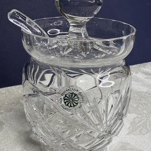 May include: A clear glass honey pot with a crystal cut design and a matching lid. The pot has a small spoon attached to the lid. The bottom of the pot has a green sticker with the text "Galway Crystal, Made in Ireland, 0123456789340".