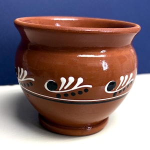 May include: A terracotta-colored clay pot with a rounded body and a flared rim. The exterior features a hand-painted horizontal band consisting of white leaf-like motifs and small black dots, set above a thin black line encircling the base.
