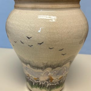 May include: Cream-colored ceramic vase with a crackle glaze. The vase features a painted landscape with birds in flight. The base of the vase has a painted landscape with green grass, blue and white clouds, and touches of yellow.