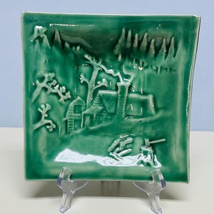 May include: A square, ceramic decorative plate with a raised relief design of a cabin in a forest scene. The plate is a solid, glossy, teal green color. The cabin has a chimney, and there are trees and mountains in the background. The plate is displayed on a clear plastic stand.