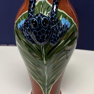 May include: A ceramic vase with a frog design. The vase is primarily brown with a green leaf design and a blue frog. The frog has black spots and blue accents. The rim of the vase is blue.
