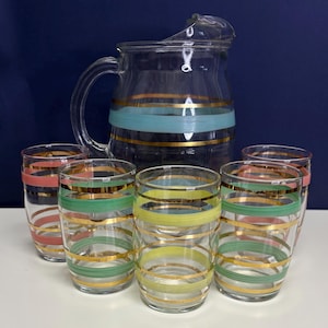 May include: A vintage glass pitcher and matching glasses set. The pitcher and glasses are clear with horizontal bands of gold, teal, pink, and yellow. The set is perfect for serving beverages.