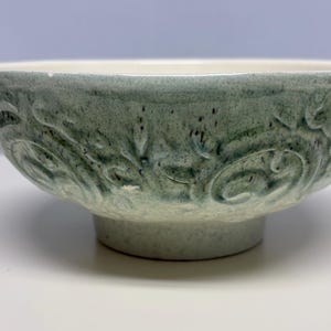 May include: A ceramic bowl with a pedestal base. The bowl is a muted green color with a textured surface and a white interior. The exterior features a repeating floral pattern. The bowl is likely used for serving food or as a decorative item.