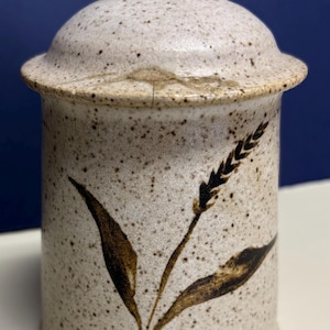 May include: A cylindrical ceramic canister with a matching lid, featuring a speckled beige finish and a dark brown painted wheat stalk design. The base of the canister displays a small, circular, embossed spiral mark in the center.