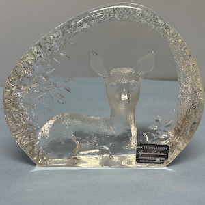 May include: A clear lead crystal sculpture featuring a frosted deer resting within a curved, textured frame. A black label on the lower right reads: MATS JONASSON Signature Collection HANDMADE IN SWEDEN LEAD CRYSTAL.