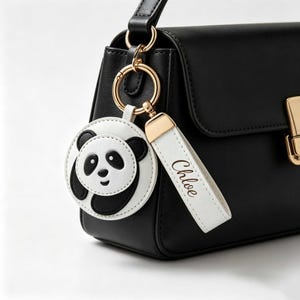 Peut inclure: Un sac à main noir doté d'un anneau en métal doré. Attaché à l'anneau se trouve un pendentif circulaire en forme de panda blanc et noir, ainsi qu'une lanière rectangulaire blanche avec le prénom Chloe imprimé en écriture brune.